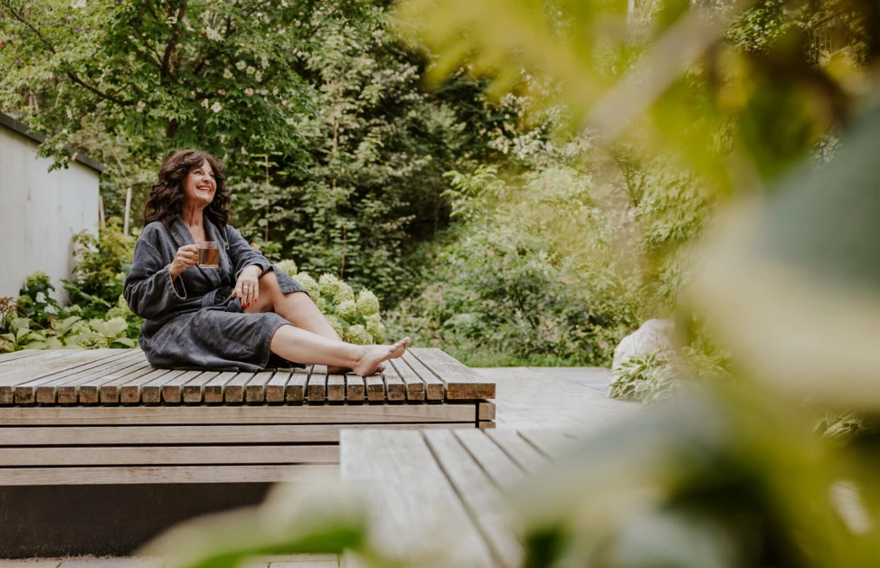 Frau in einem Bademantel sitzt entspannt auf einer Holzliege und genießt eine Tasse Tee in einer grünen Gartenlandschaft.