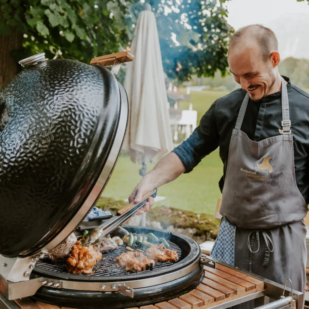 Ein Koch grillt Fleisch und Gemüse auf einem großen Grill im Freien, umgeben von einer schönen, grünen Landschaft.