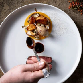 Ein Gericht mit Fleisch, Saucen und kunstvoll angerichtetem Gemüse auf einem weißen Teller, während eine Hand mit einem Löffel serviert.