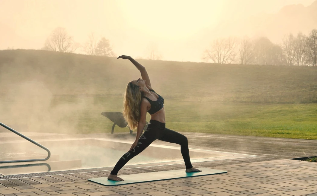 Eine Frau macht Yoga auf einer Matte bei Sonnenaufgang neben einem Pool, Hotel Klosterhof im Hintergrund.