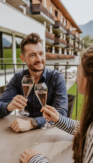 Ein Paar stößt mit Sektgläsern an, während sie auf der Terrasse des Hotel Klosterhof sitzen, umgeben von der schönen Alpenlandschaft.