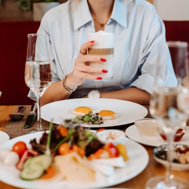 Eine lächelnde Frau mit einem Kaffee in der Hand, umgeben von einem reichhaltigen Frühstückstisch in einem eleganten Restaurant.