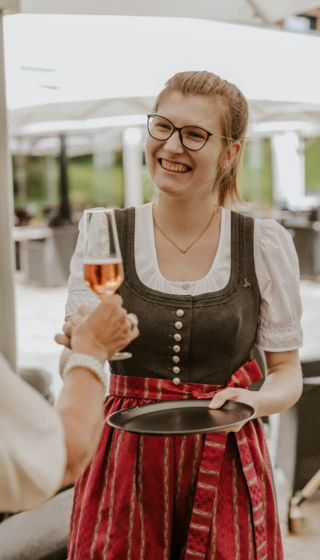 A smiling woman in traditional attire serves a drink outdoors.